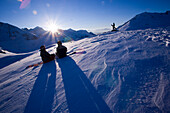Two people sitting with snowboards on slope, one person standing in background, Kuehtai, Tyrol, Austria