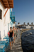 Women strolling along restaurants and bars at bank, windmills in background, Little Venice, Mykonos-Town, Mykonos, Greece