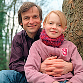 Father hugging arms around daughter, both sitting on a tree, Germany