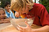 Junge mit nassen Haaren hält einen Apfel im Mund, Schüssel mit Wasser vor ihm, Kindergeburtstag