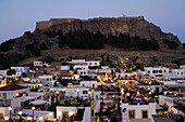 View over illuminated town in the evening to Acropolis, people sitting on terraces of restaurants, Lindos, Rhodes, Greece