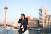 Young business woman talking on mobile phone in front of the city skyline, television tower, Zollhof, Media Harbour, architecture of Frank O.Gehry, Düsseldorf, state capital of NRW, North-Rhine-Westphalia, Germany