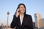 Young business woman talking on mobile phone, in front of city skyline, television tower, Zollhof, Media Harbour, architecture of Frank O.Gehry, Düsseldorf, state capital of NRW, North-Rhine-Westphalia, Germany