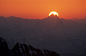 Sonnenscheibe hinter dem Guffert mit Felsgipfeln mit Gipfelkreuz im Vordergrund, Wilder Kaiser, Tirol, Österreich