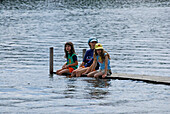 three girls at landing stage with legs in water, lake Staffelsee, Upper Bavaria, Bavaria, Germany