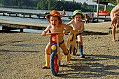 two boys with sunhat on juvenile bicycle, beach of lake Hartsee, Chiemgau, Upper Bavaria, Bavaria, Germany