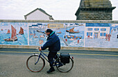 Retired marin cycling by a road with a mural painting wall and Eckmül lighthouse in background. Penmarc h. Finistere. Brittany. France