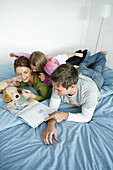 Junge Familie liegt auf einem Bett und liest ein Buch, München, Deutschland