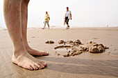 Kid s feet standing by water hole in sand, background: people walking along the beach.