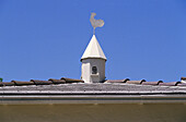White rooster weathervane atop small white towerlike feature on tile roof, against blue sky. Los Angeles. California. USA