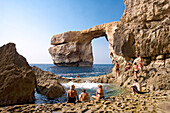 Menschen sitzen am Meer auf Felsen, Felsbogen im Hintergrund, Gozo, Malta, Europa