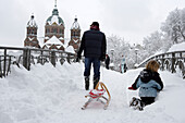 Menschen mit Schlitten gehen an einem Wintertag über eine Brücke, im Hintergrund St. Lukas, München, Bayern, Deutschland