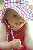 18 month old girl outside in big floppy hat, clasping her hands together in excitement.