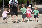The back and legs of a man walking down a street holding four small childrens hands, all walking in a row