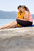 Two young women sitting a lake Walchensee while listening to music about earphones, Bavaria, Germany