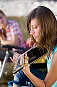 A young woman sitting on the banks of the river Isar in the evening playing the guitar, Munich, Bavaria, Germany