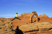 Female hiker leaping from rock to rock with Delicate Arch in background, Arches National Park near Moab, Utah, USA