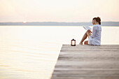 Frau sitzt auf einem Steg am Starnberger See und liest ein Buch, Ambach, Bayern, Deutschland