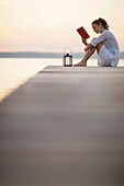 Frau sitzt auf einem Steg am Starnberger See und liest ein Buch, Ambach, Bayern, Deutschland
