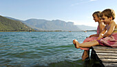 Zwei Mädchen sitzen auf einem Steg am See im Sonnenlicht, Kalterer See, Südtirol, Italien, Europa