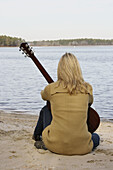 Blond woman holding a guitar sitting down by the lake looking at the lake