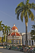 People sitting on a bench in Parque Marti in front of a red roofed gazebo in front of the Red roofed Palacio de Gobierno .