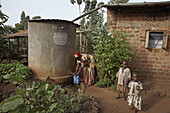 UGANDA  In the home of Najjemba Teopista, Kasaayi village, Kayunga District  Teopista taking harvested rainwater from a tank beside house, while Kevin and Maria stand by