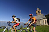 Two mountain bikers near Sankt Kathrein church, Hafling, Trentino-Alto Adige/Südtirol, Italy
