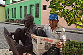 Man reading a newspaper next to a reading-man-sculpture, Plaza del Carmen, Camaguey, Camaguey, Cuba, West Indies