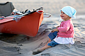 A one year old girl sitting on the beach next to a sea kayak, Punta Conejo, Baja California Sur, Mexico