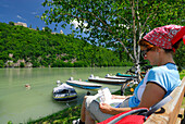 Frau sitzt auf Bank am Ufer der Donau und liest, Schloss Neuhaus im Hintergrund, Donauradweg Passau Wien, Oberösterreich, Österreich