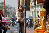 Mädchen mit Handy vor Devotionalien Handel, Sitzender und stehende Buddhas stehen auf dem Bürgersteig, Altstadt, Bamrung Muang, Bangkok, Thailand, Asien