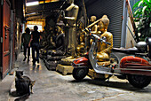 Devotionalien Handel, Sitzende und stehende Buddhas stehen auf dem Bügersteig in einer Hinterhofgasse mit Motorroller, Katzen und Personen, Bamrung Muang, Altstadt, Bangkok, Thailand, Asien