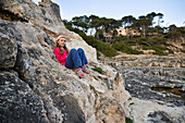 Mädchen sitzt auf einem Felsen an der Küste, Cala Santanyi, Mallorca, Balearen, Mittelmeer, Spanien, Europa