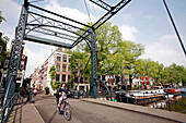 Woman Riding A Dutch Bike On A Bridge, Houseboat At The Quay Of A Canal And Facade Of A Traditional House, Amsterdam, Netherlands, Holland