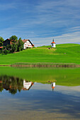 Reflection of upper Bavarian traditional farmhouse and chapel on lake, East Allgaeu, Bavaria, Germany