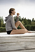 Junge Frau mit einem Apfel sitzt auf einem Steg am Starnberger See, Bayern, Deutschland