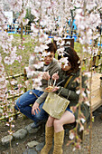 A young Japanese couple sitting on a bench eating some food with chop sticks while surrounded by hanging branches filled with pink cherry blossoms in Maruyama koen in Kyoto city.