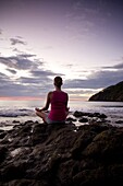 Young woman meditating on rocks at sunset in front of ocean surf at Playas del Coco, Costa Rica