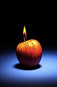 An apple doubling as a candle, by igniting the stem, on a shadowy blue and black background