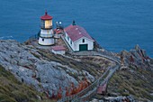USA, California, Marin County, Point Reyes National Seashore, old lighthouse on the left, modern light on the right, perched on a point of Point Reyes conglomerate rock with red lichen, staircase and resting platform for visitors, dusk