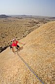 Frau klettert an roter Felsplatte über Savanne, Große Spitzkoppe, Namibia
