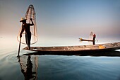 An Intha fisherman with traditional fish trap uses an unusual leg-rowing technique to propel his flat-bottomed boat across the lake while standing.