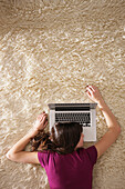 Junge Frau liegt mit einem Laptop auf einem Teppich, München, Bayern, Deutschland