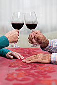 Hände von zwei alten Menschen, sie prosten sich mit einem Glas Rotwein zu