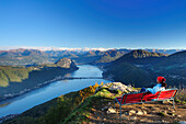 Frau sitzt auf roter Bank und blickt auf Luganer See, Tessiner Alpen im Hintergrund, Blick vom  Monte San Giorgio, UNESCO Weltkulturerbe Monte San Giorgio, Luganer See, Tessiner Alpen, Tessin, Schweiz, Europa