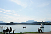Badegäste sitzen am Ufer des Faaker See mit Ruderboot und Segelboot, Faaker See, Kärnten, Österreich, Europa