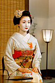 Portrait of a Maiko (Apprentice Geisha) in a Kimono (Obede), Wearing a Chignon High on Her Head in the Form of a Peach (Wareshinobu) Adorned with a Red Silk Ribbon(Kanoko), a Decorated Hairpin (Kanzashi) and a Bouquet of Flowers (Hanakanzashi) and Made Up