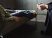 Young woman lying on couch, therapist leaning toward her with hands clasped, cropped