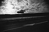 Sign post on road in front of sky with clouds, b&w